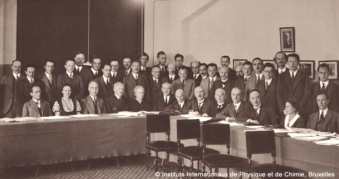 Alle Teilnehmer des Solvay-Kongresses, einige sitzend an einem Tisch, andere stehend dahinter