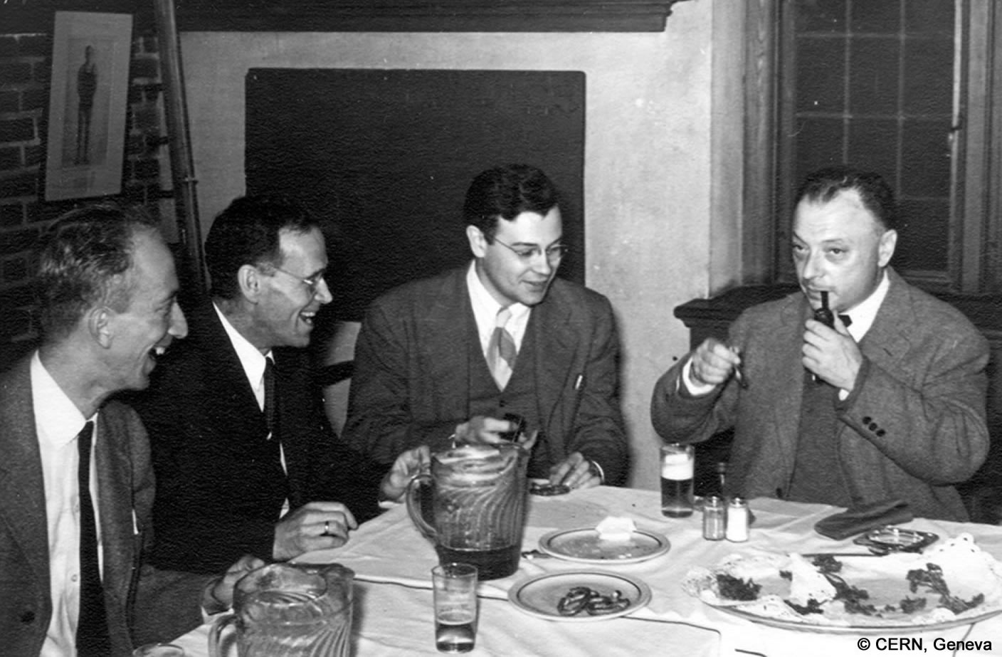 Wolfgang Pauli sitzt mit drei Männern an einem Tisch und ist dabei, sich eine Pfeife anzuzünden. Es stehen leere Teller und Gläser auf dem Tisch