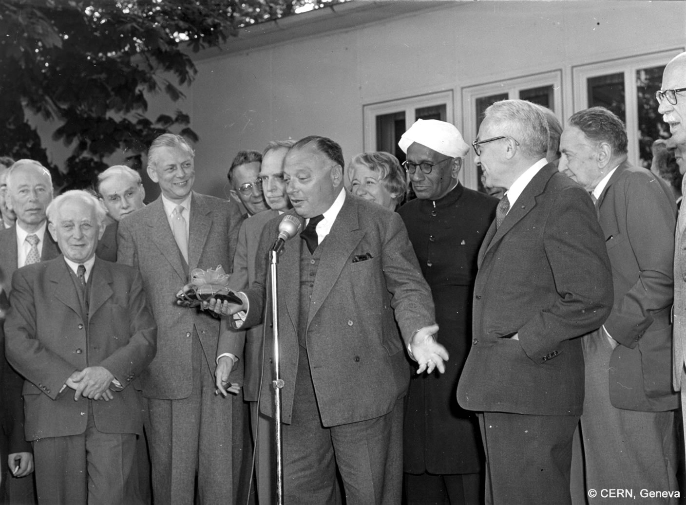 Wolfgang Pauli steht sprechend vor einem Mikrofon, hinter ihm stehen Männer