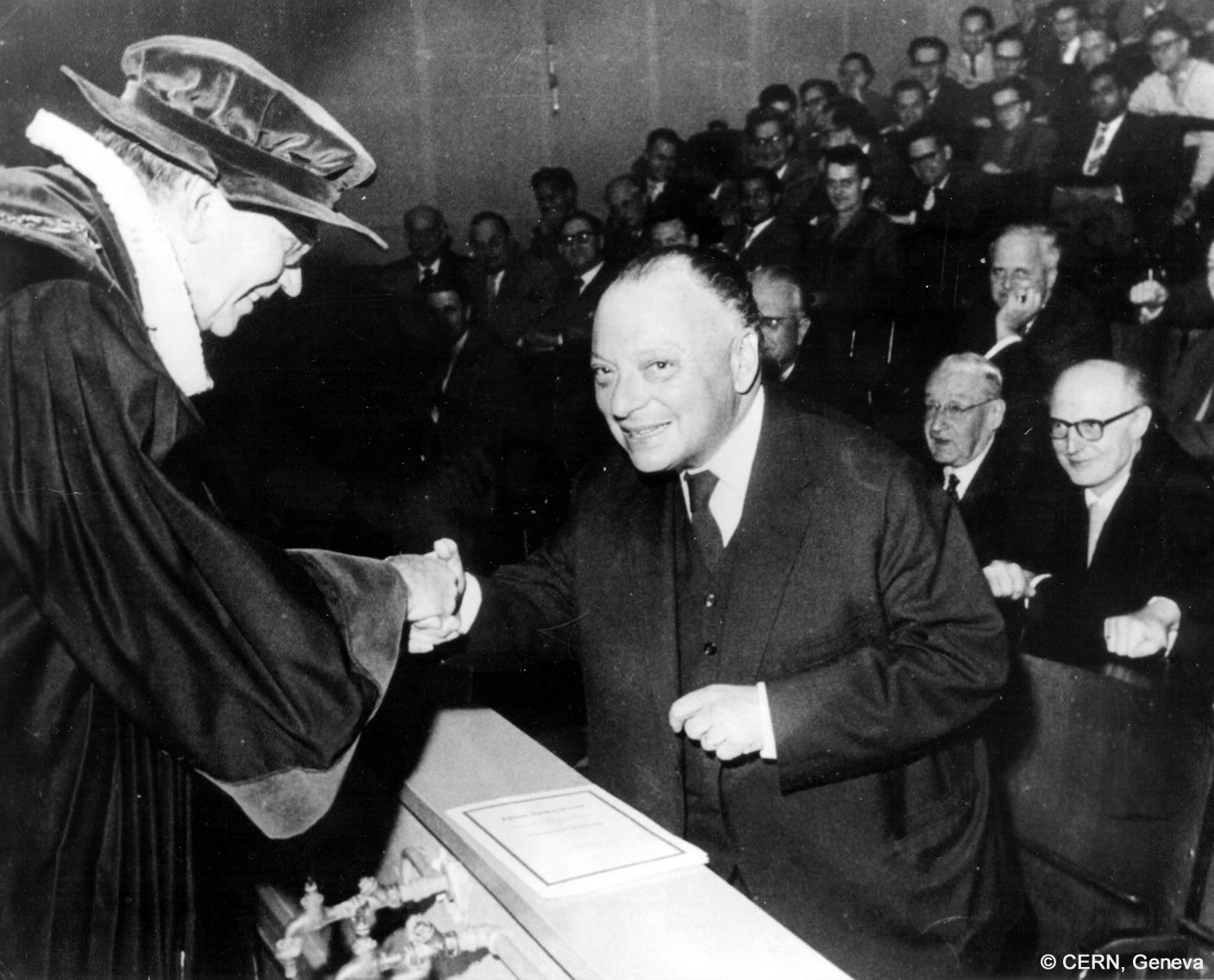 Wolfgang Pauli shakes a man's hand when he is awarded an honorary doctorate. In the background there are numerous sitting men.