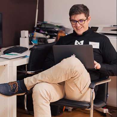 Ein junger Mann sitzt lächelnd auf einem Stuhl vor dem Notebook