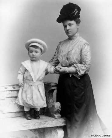 Wolfgang Pauli als kleines Kind, stehend auf einer Bank neben seiner Mutter. Sie hält ihn an der Hand und am Rücken.